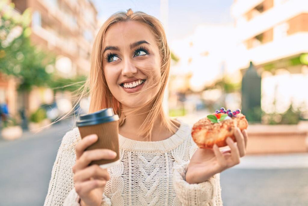 Jeune fille blonde souriante heureuse de prendre le petit déjeuner debout à la ville. - Photo, image
