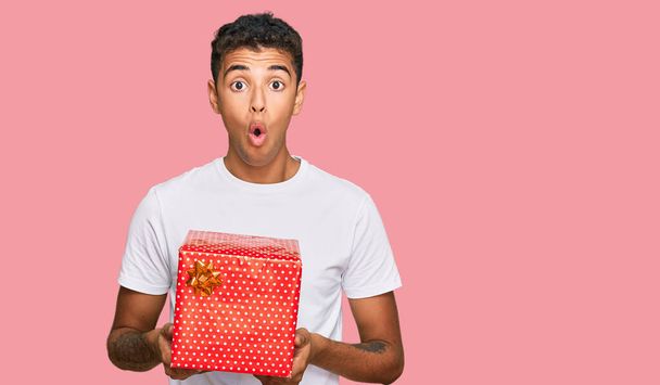 Νεαρός όμορφος Αφροαμερικάνος που κρατάει το δώρο φοβισμένος και έκπληκτος με ανοιχτό το στόμα για έκπληξη, δυσπιστία πρόσωπο  - Φωτογραφία, εικόνα