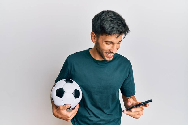 Mladý pohledný muž drží míč při pohledu na smartphone bet app usměvavý a smát se nahlas, protože legrační šílený vtip.  - Fotografie, Obrázek