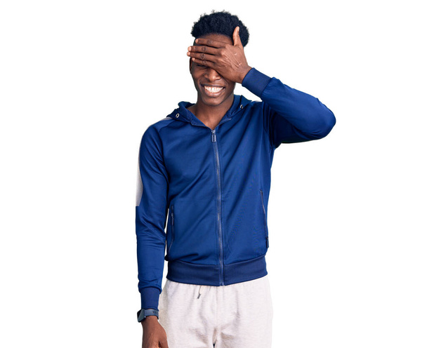Giovane uomo afroamericano che indossa abbigliamento sportivo sorridente e ridente con mano sul viso che copre gli occhi per la sorpresa. concetto cieco.  - Foto, immagini