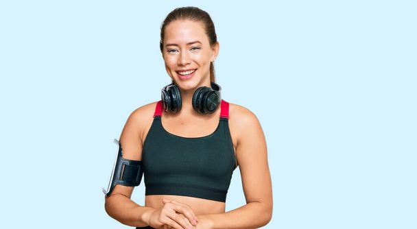 Beautiful blonde woman wearing gym clothes and using headphones with hands together and crossed fingers smiling relaxed and cheerful. success and optimistic  - Photo, Image