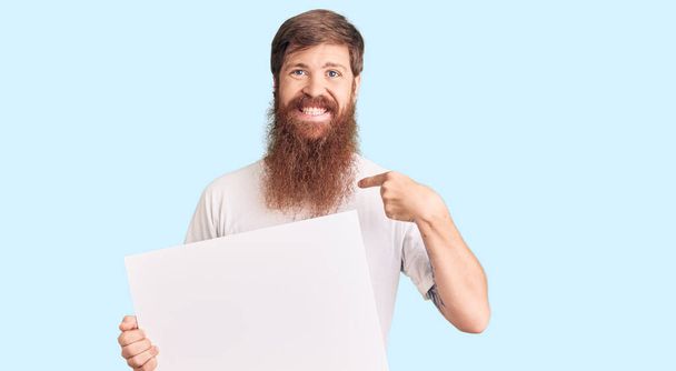 Красивый молодой рыжий мужчина с длинной бородой держит пустой баннер указывая пальцем на одного улыбающегося счастливого и гордого  - Фото, изображение