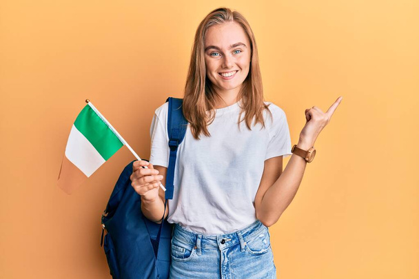Linda loira troca estudante segurando bandeira irlandesa sorrindo feliz apontando com a mão e o dedo para o lado  - Foto, Imagem