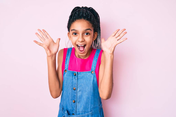 Młody Afroamerykanin dziewczyna dziecko z warkoczami noszenie ubrań na różowym tle świętując szaleństwo i zdumiony sukcesem z ramionami podniesione i otwarte oczy krzycząc podekscytowany. koncepcja zwycięzcy  - Zdjęcie, obraz