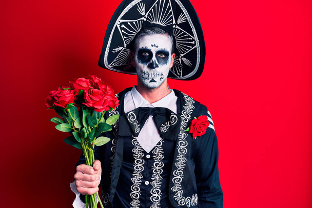 Молодой человек, одетый в мексиканский день мертвого костюма, держит в руках розы, думая о своем поведении и трезвом выражении лица.  - Фото, изображение