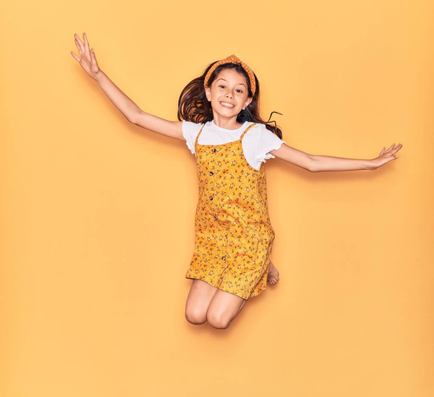 Imádnivaló spanyol gyereklány alkalmi ruhában és diadém mosollyal. Ugrás mosollyal az arcon és a karok nyitott izolált sárga háttér - Fotó, kép