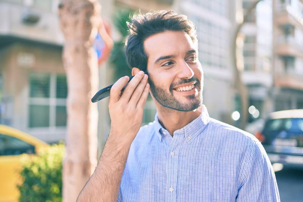 Genç İspanyol adam gülümsüyor. Şehirdeki akıllı telefonu kullanarak mutlu bir sesli mesaj dinliyor.. - Fotoğraf, Görsel