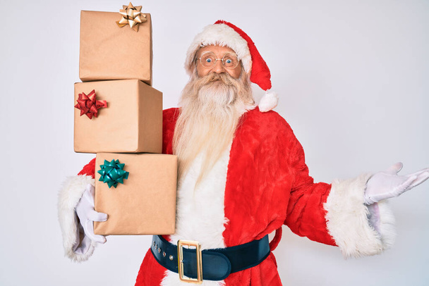 Staruszek z siwymi włosami i długą brodą w kostiumie Świętego Mikołaja z prezentami świętującymi zwycięstwo szczęśliwym uśmiechem i zwycięską ekspresją z podniesionymi rękami  - Zdjęcie, obraz