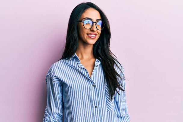 Belle femme hispanique portant des vêtements décontractés et des lunettes regardant vers le côté, pose de profil relax avec visage naturel et sourire confiant.  - Photo, image