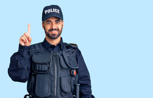Junger hispanischer Mann in Polizeiuniform zeigt mit Finger Nummer eins nach oben und lächelt dabei selbstbewusst und glücklich.  - Foto, Bild