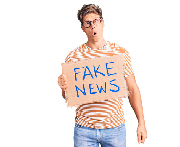 Young handsome man holding fake news banner scared and amazed with open mouth for surprise, disbelief face  - Foto, Imagem