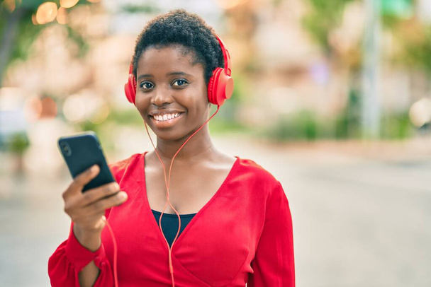 Młoda Afroamerykanka uśmiecha się szczęśliwa używając smartfona i słuchawek w mieście. - Zdjęcie, obraz