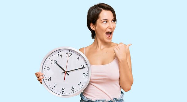 Young brunette woman with short hair holding big clock pointing thumb up to the side smiling happy with open mouth  - Photo, Image