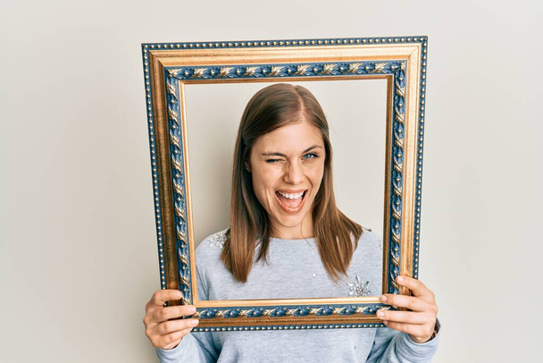 Beautiful caucasian woman holding empty frame winking looking at the camera with sexy expression, cheerful and happy face.  - Photo, Image