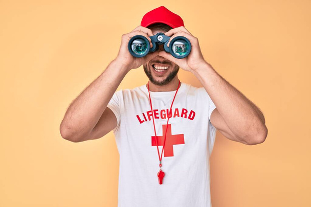 Jovem caucasiano vestindo camiseta salva-vidas usando binóculos piscando olhando para a câmera com expressão sexy, rosto alegre e feliz.  - Foto, Imagem
