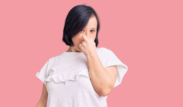 Brunette woman with down syndrome wearing casual white tshirt smelling something stinky and disgusting, intolerable smell, holding breath with fingers on nose. bad smell  - Foto, immagini