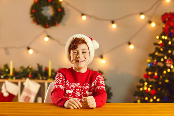 Happy boy in traditional Christmas jumper and Santa cap sitting at table and smiling - Valokuva, kuva