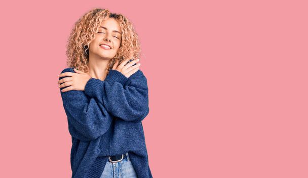 Mujer rubia joven con el pelo rizado usando suéter de invierno casual abrazándose feliz y positivo, sonriendo confiado. amor propio y cuidado personal  - Foto, Imagen