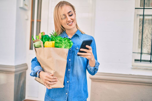 Молодая блондинка улыбается счастливой, используя смартфон и держа в руках бумажный пакет в городе. - Фото, изображение