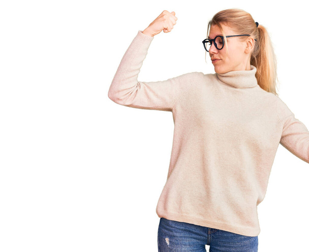 Young beautiful blonde woman wearing turtleneck sweater and glasses showing arms muscles smiling proud. fitness concept.  - Fotó, kép
