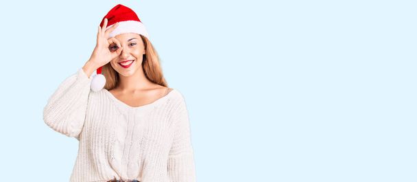 Kaunis nuori nainen yllään joulu hattu hymyillen onnellinen tekee ok merkki käsin silmään katselee sormien läpi  - Valokuva, kuva