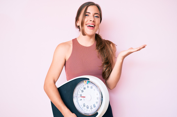 Beautiful young caucasian woman holding weight machine to balance weight loss celebrating victory with happy smile and winner expression with raised hands  - Photo, Image