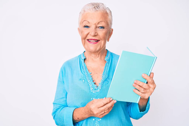 Старшая красивая женщина с голубыми глазами и седыми волосами читает книгу, выглядящую позитивно и счастливо стоя и улыбаясь с уверенной улыбкой, показывая зубы  - Фото, изображение