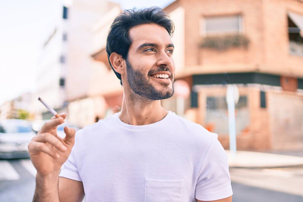 Young hispanic man smiling happy holding cigarette walking at the city. - Fotoğraf, Görsel