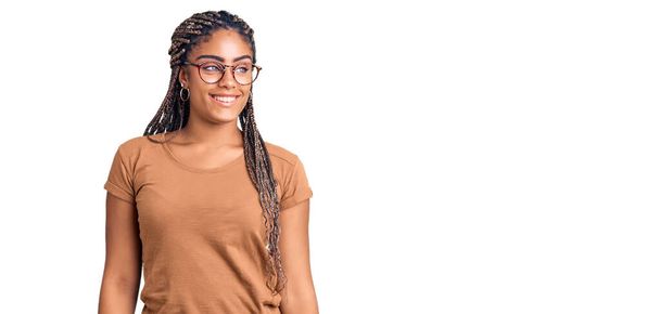 Young african american woman with braids wearing casual clothes and glasses looking away to side with smile on face, natural expression. laughing confident.  - Photo, Image