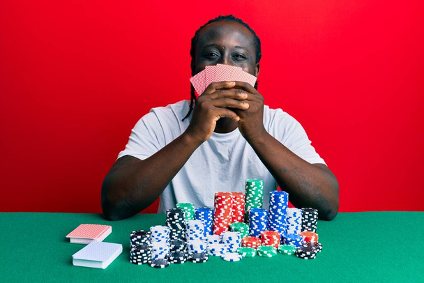 Jóképű fekete férfi játszik szerencsejáték póker fedő arc kártyák mosolygó boldog és hűvös mosollyal az arcán. fogak megjelenítése.  - Fotó, kép