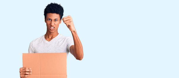Νεαρός Αφροαμερικάνος κρατώντας πανό από χαρτόνι ενοχλημένος και απογοητευμένος φωνάζοντας από θυμό, φωνάζοντας τρελός από θυμό και σηκωμένος στο χέρι  - Φωτογραφία, εικόνα