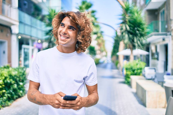 Νεαρός Ισπανόφωνος χαμογελώντας χαρούμενος χρησιμοποιώντας smartphone στο δρόμο της πόλης - Φωτογραφία, εικόνα