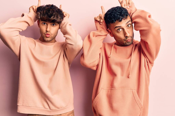 Young gay couple wearing casual clothes doing funny gesture with finger over head as bull horns  - 写真・画像