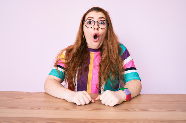 Mladá zrzka žena sedí na stole s brýlemi vystrašená a užaslá s otevřenými ústy pro překvapení, nevěřící tvář  - Fotografie, Obrázek