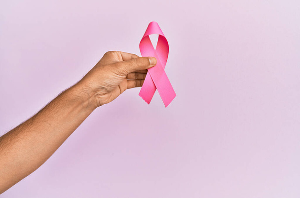 Hand of young hispanic man holding pink breast cancer ribbon over isolated background. - Foto, Imagen