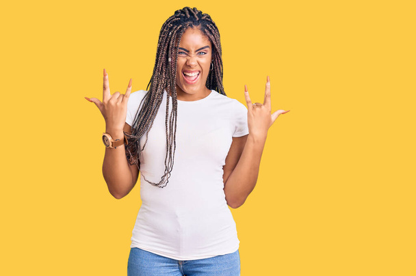 Junge afrikanisch-amerikanische Frau mit Zöpfen in lässiger Kleidung schreit mit verrücktem Gesichtsausdruck und macht Rock-Symbol mit erhobenen Händen. Musikstar. Schweres Konzept.  - Foto, Bild