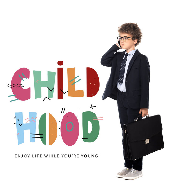 curly boy in suit and glasses holding briefcase while talking on smartphone near childhood, enjoy life while you are young lettering on white - Zdjęcie, obraz