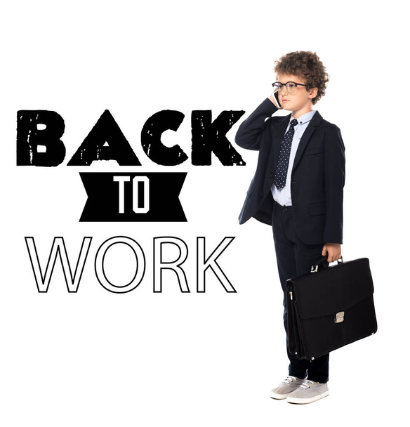 curly boy in suit and glasses holding briefcase while talking on smartphone near back to work lettering on white - Photo, Image