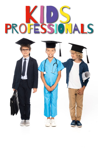 niños con gorras de graduación vestidos con disfraces de diferentes profesiones de pie cerca de los niños profesionales de letras en blanco  - Foto, imagen
