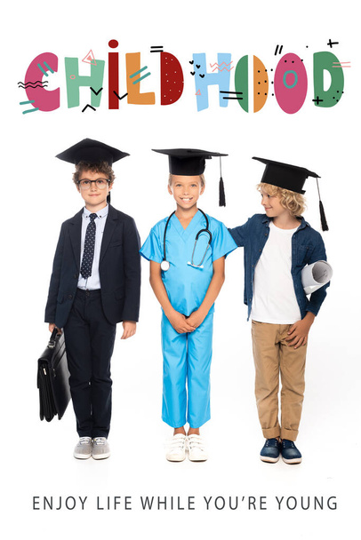 children in graduation caps dressed in costumes of different professions standing near childhood lettering on white  - Valokuva, kuva