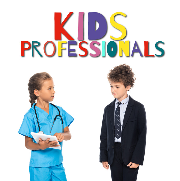 kind in kostuum van arts met behulp van digitale tablet in de buurt krullende jongen in formele slijtage en kinderen professionals belettering op wit  - Foto, afbeelding