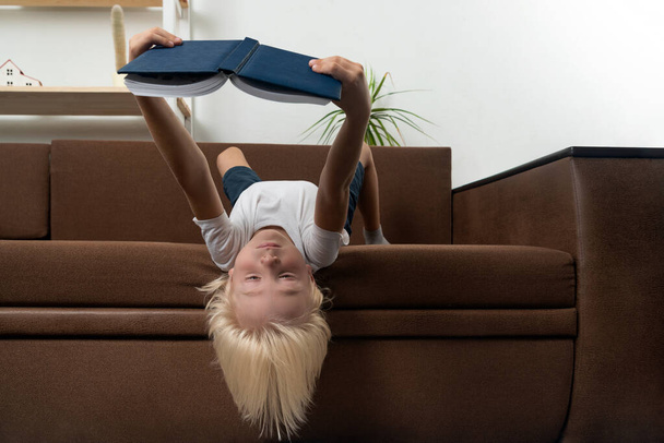 Boy lies on sofa upside down and reads book. Child loves to read. Homeschooling. - Photo, image