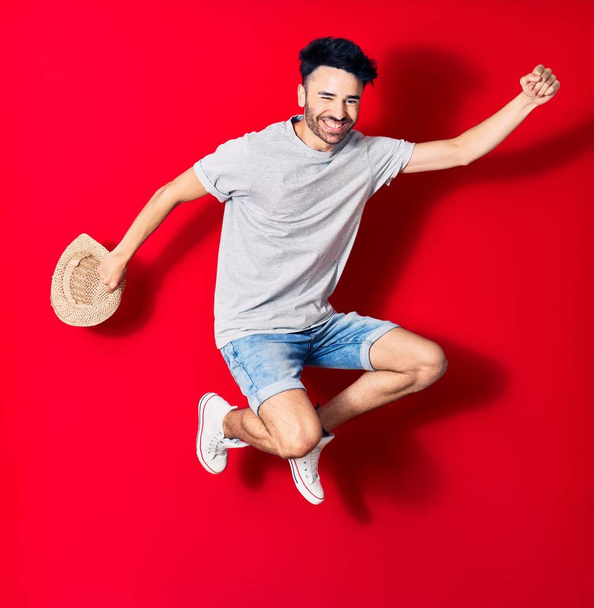 Junger gut aussehender hispanischer Mann in lässiger Kleidung, der glücklich lächelt. Mit einem Lächeln im Gesicht hüpfend mit Sommermütze feiernd mit erhobener Faust über isoliertem rotem Hintergrund. - Foto, Bild