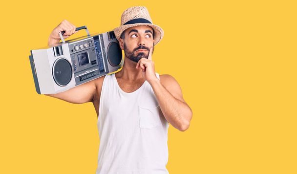 Joven hombre hispano sosteniendo boombox, escuchando música cara seria pensando en la pregunta con la mano en la barbilla, pensativo acerca de la idea confusa  - Foto, Imagen