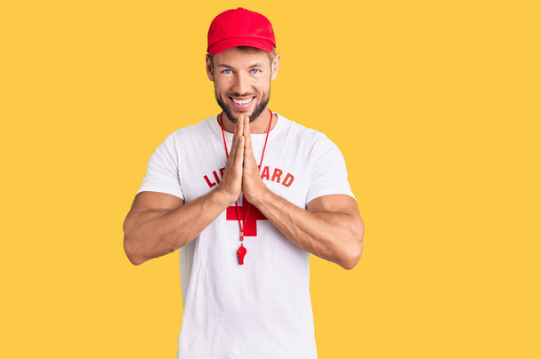 Young caucasian man wearing lifeguard t shirt holding whistle praying with hands together asking for forgiveness smiling confident.  - Photo, Image