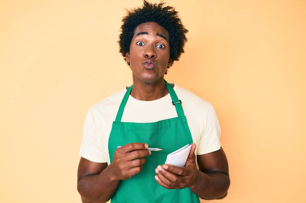 Handsome african american man with afro hair wearing waiter apron taking order looking at the camera blowing a kiss being lovely and sexy. love expression.  - Photo, Image