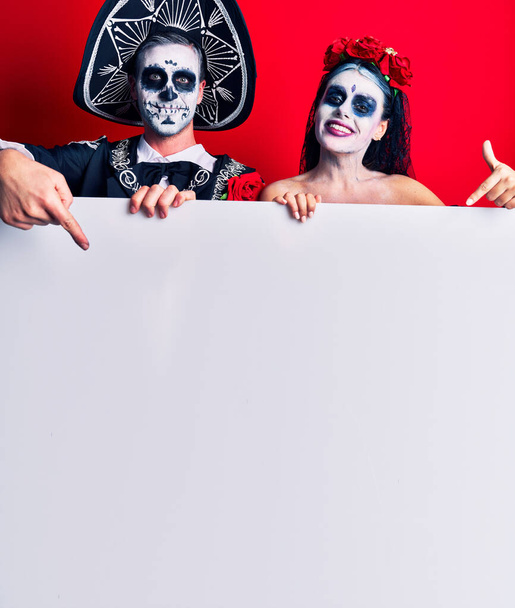 Junges Paar im mexikanischen Totenkostüm mit leerem Transparent, glücklich lächelnd und mit Hand und Finger zeigend  - Foto, Bild