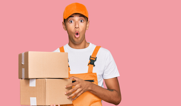 Jovem afro-americano bonito segurando pacote de entrega assustado e surpreso com a boca aberta para surpresa, rosto descrença  - Foto, Imagem