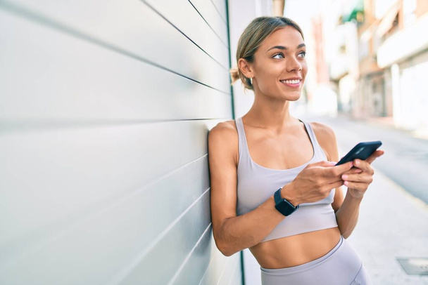 スマートフォンを使用して屋外でスポーツウェアのトレーニングを身に着けている若い白人フィットネス女性 - 写真・画像