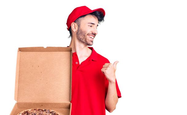 Młody Hiszpan trzyma pudełko z pizzą skierowaną kciukiem w górę, uśmiechnięty szczęśliwy z otwartymi ustami.  - Zdjęcie, obraz
